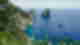 Green shubbery and grey cliffs with villas built in to the side overlooking sapphire ocean with small cruise ships