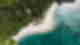 Deep turquoise ocean with some boats that curves around white sand beach and green jungle with a few houses, aerial shot